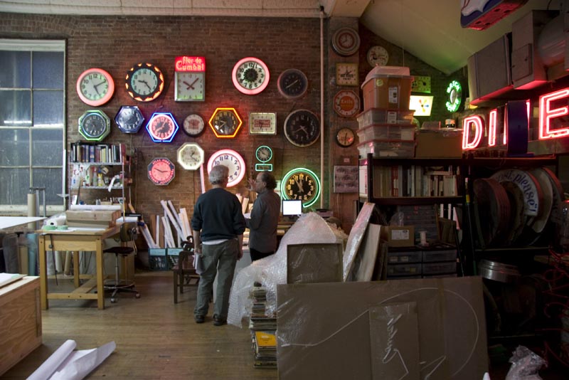 A view of the Let There Be Neon showroom with lit neon signs on the walls and two people talking