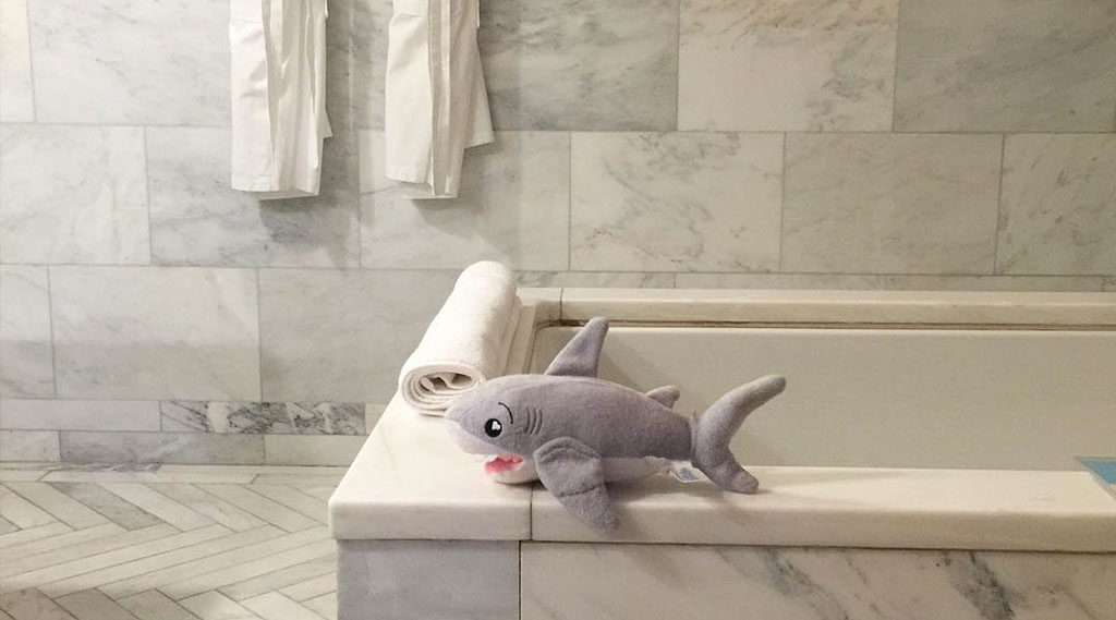 A bathroom bathtub with a stuffed shark on the edge, marble tiles, and white towels.