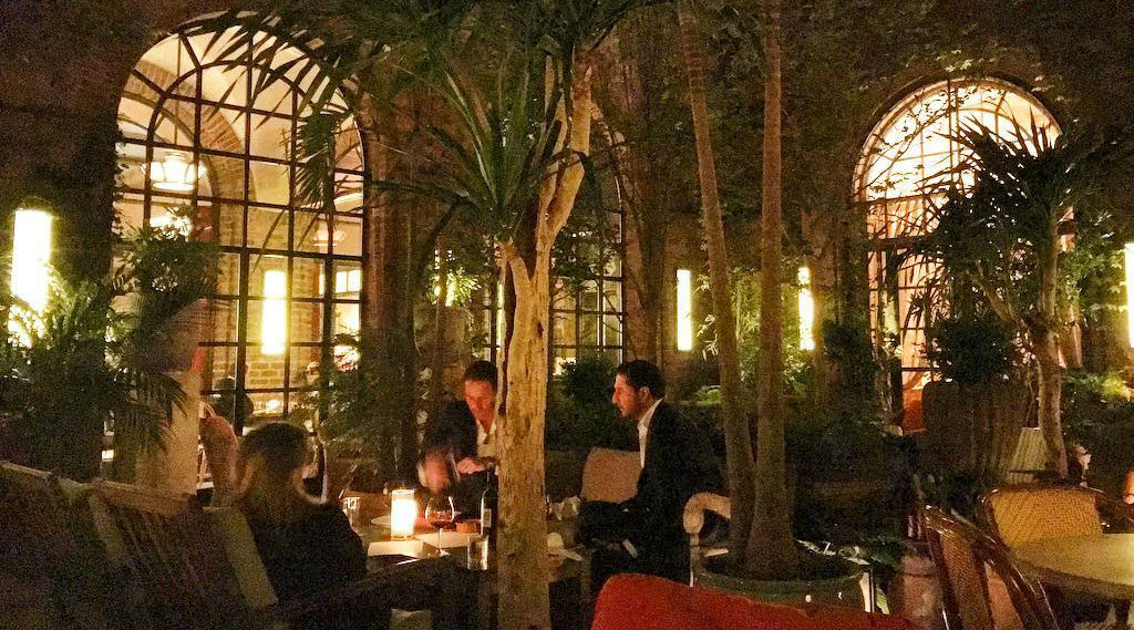 A dimly lit indoor dining area with large windows, greenery, and people dining at tables.