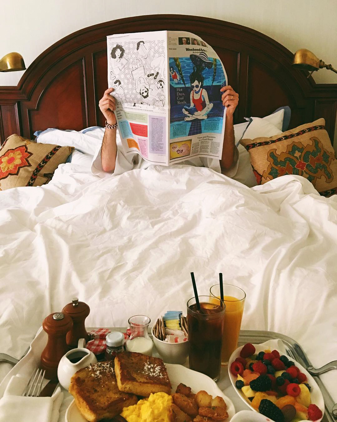 A person reading a newspaper in bed with a breakfast tray in front of them.
