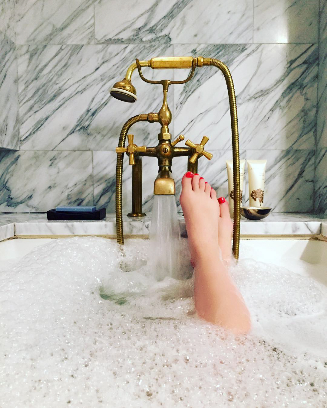 A person's feet in a bubble bath with a brass faucet and marble wall