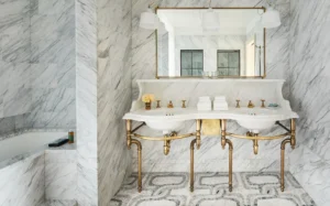 Looking straight on at the double sink vanity with wide mirror and sconces. Featuring marble finishes and an alcove soaking tub on the left.