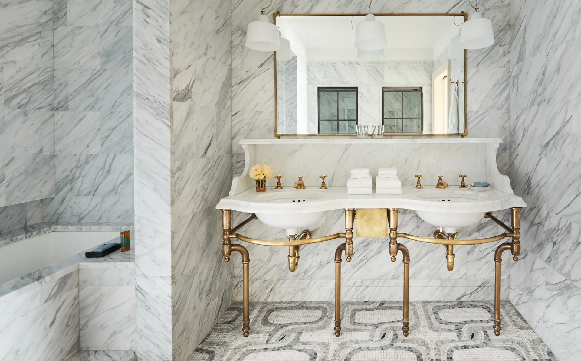 Looking straight on at the double sink vanity with wide mirror and sconces. Featuring marble finishes and an alcove soaking tub on the left.