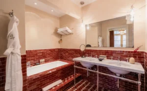 A view of red moroccan tile surrounding a soaking tub in a courtyard suite bathroom. A large mirror and marble double vanity is right next to the tub on the right.