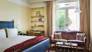 Looking over the bed at the large window and loveseat. Shelves and a desk appear left of the loveseat and window.