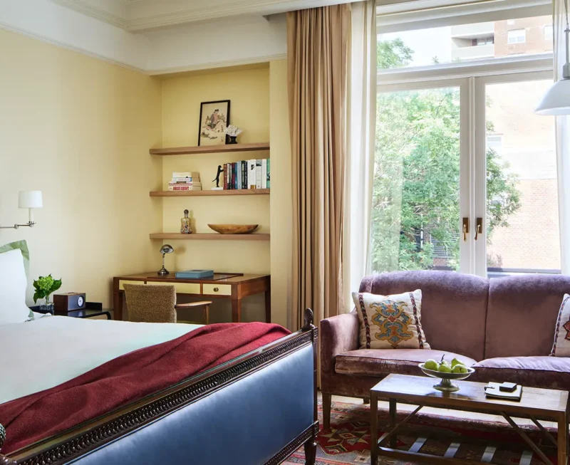 Looking over the bed at the large window and loveseat. Shelves and a desk appear left of the loveseat and window.