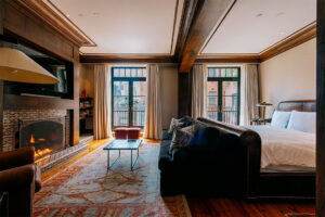 A bedroom appointed with wooden beams a fireplace and a king sized bed with large floor to ceiling windows