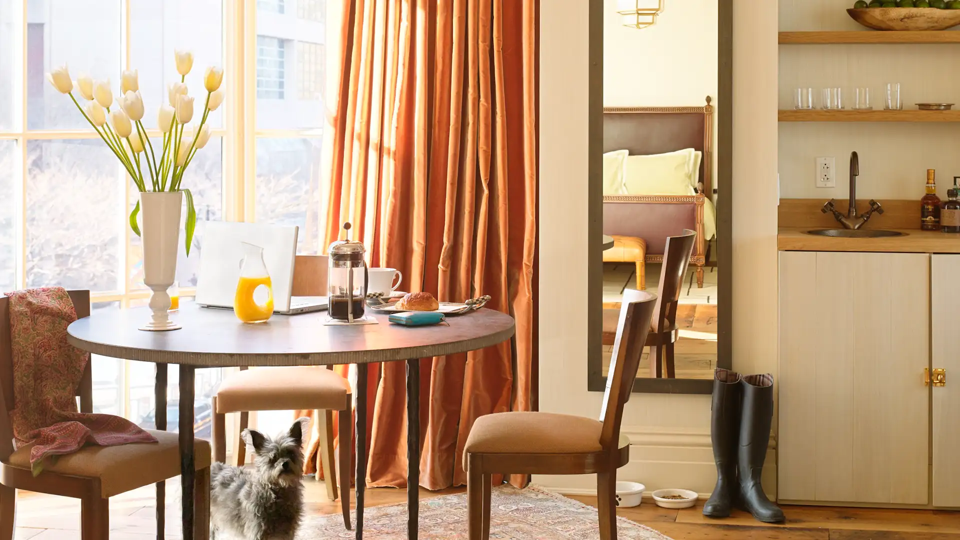 Two Bedroom Corner Suite Dining Table and chairs. A small dog peaking out from under the table. Laptop and breakfast is on top of the table, highlighted by natural light from the large window.